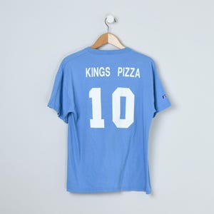 Puede incluir: Camiseta azul claro con el texto "KINGS PIZZA" y el n&uacute;mero "10" en blanco. La camiseta est&aacute; colgada de una percha de madera. El logotipo de Champion es visible en la manga. Fondo blanco.