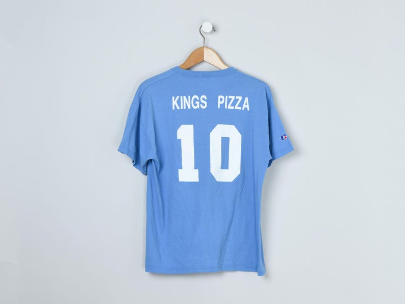 Puede incluir: Camiseta azul claro con el texto "KINGS PIZZA" y el n&uacute;mero "10" en blanco. La camiseta est&aacute; colgada de una percha de madera. El logotipo de Champion es visible en la manga. Fondo blanco.
