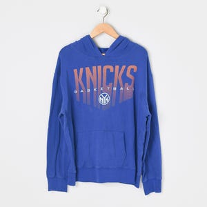 May include: A royal blue long-sleeve hooded sweatshirt featuring the text KNICKS BASKETBALL in a gradient orange font across the chest. Below the text is a circular white logo containing the letters NYK. The hoodie has a front kangaroo pocket.