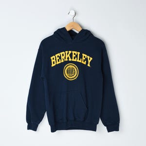 Może przedstawiać: Granatowa bluza z kapturem z żółtym napisem "BERKELEY" wygiętym łukiem na piersi. Poniżej tekstu znajduje się żółty okrągły emblemat. Bluza ma przednią kieszeń i wisi na drewnianym wieszaku.