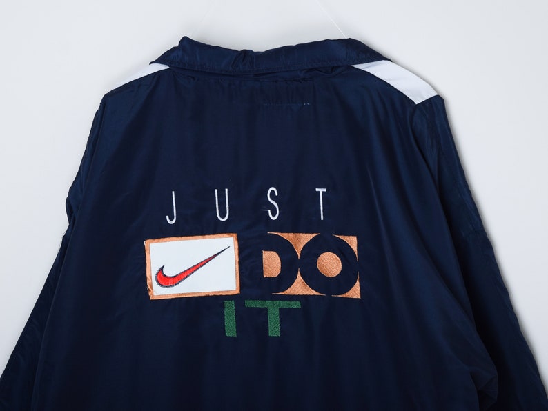 May include: A navy blue windbreaker jacket with white stripes on the shoulders and a white "JUST DO IT" logo on the back. The logo is in a box with a red Nike swoosh.