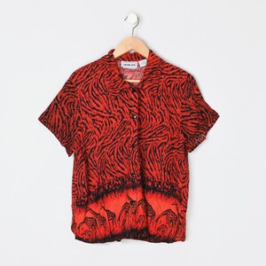 May include: A short-sleeved button-down shirt featuring a vibrant red and black tiger stripe pattern on the upper portion and a black and red graphic print of giraffes along the bottom hem, complete with a classic collar and front button closure.