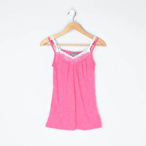 May include: A vibrant pink camisole top with white spaghetti straps and white lace trim on the neckline. The top is displayed on a wooden hanger against a white backdrop. The camisole is a solid colour.