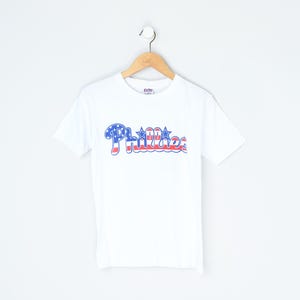 May include: White t-shirt with the word "Phillies" in red, white, and blue, reminiscent of the American flag. The shirt is hanging on a wooden hanger against a white background. The t-shirt is a classic crew neck style.