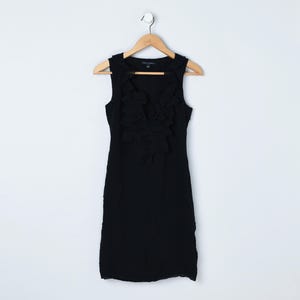 May include: A sleeveless black dress with a ruffled front detail, hanging on a wooden hanger against a white background. The dress has a fitted silhouette and appears to be made of a textured fabric.