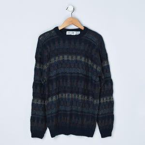 May include: A navy blue sweater with a geometric pattern in shades of blue, gray, and tan. The sweater is hanging on a wooden hanger against a white background. The sweater has a crew neck and long sleeves.