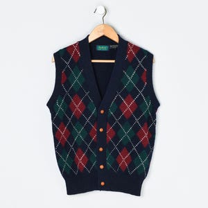 May include: A navy blue V-neck sweater vest featuring a repeating argyle pattern in dark red and forest green with thin white intersecting lines. The garment has a button-down front with five light brown buttons and a ribbed hem at the bottom.