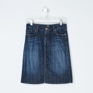 May include: A dark wash denim skirt hangs from a clear hanger against a white background. The skirt has a classic five-pocket design, a button and zipper closure, and a knee-length hemline. The denim has subtle fading.