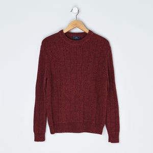 May include: A burgundy crew neck sweater hangs on a wooden hanger against a white background. The sweater has a textured knit pattern and ribbed cuffs and hem. The garment is made of a soft, warm material, perfect for cooler weather.