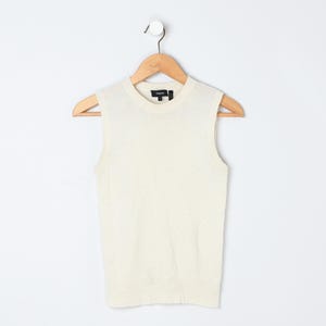 Puede incluir: Top sin mangas de color crema colgado de una percha de madera. El top tiene cuello redondo y una textura acanalada. El nombre de la marca es visible en el escote. La prenda se muestra sobre un fondo blanco liso.