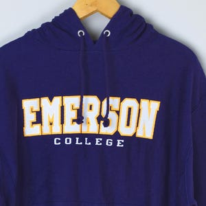 May include: A navy blue hooded sweatshirt with the text "EMERSON COLLEGE" in white and gold lettering. The sweatshirt is hanging on a wooden hanger. The drawstring hood is visible.