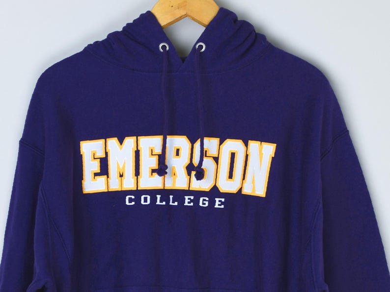 May include: A navy blue hooded sweatshirt with the text "EMERSON COLLEGE" in white and gold lettering. The sweatshirt is hanging on a wooden hanger. The drawstring hood is visible.
