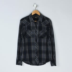 Puede incluir: Camisa a cuadros negros y grises con mangas largas. La camisa tiene dos bolsillos en el pecho y un cuello. La camisa está colgada en una percha de madera sobre un fondo blanco. La marca "HELIX" es visible.