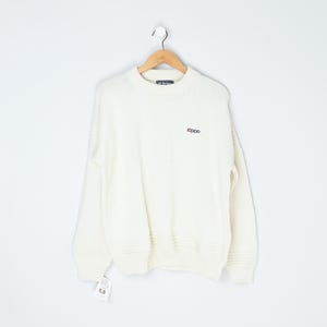 May include: Cream-colored sweater with the "zippo" logo embroidered in black. The sweater is hanging on a wooden hanger against a white background. The sweater has a ribbed hem and cuffs. A price tag is attached to the bottom.
