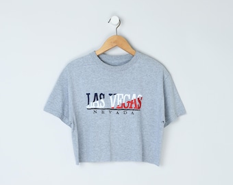 May include: A gray cropped t-shirt with the words "LAS VEGAS NEVADA" in a red, white, and blue design. The shirt is hanging on a wooden hanger against a white background. The t-shirt has short sleeves.