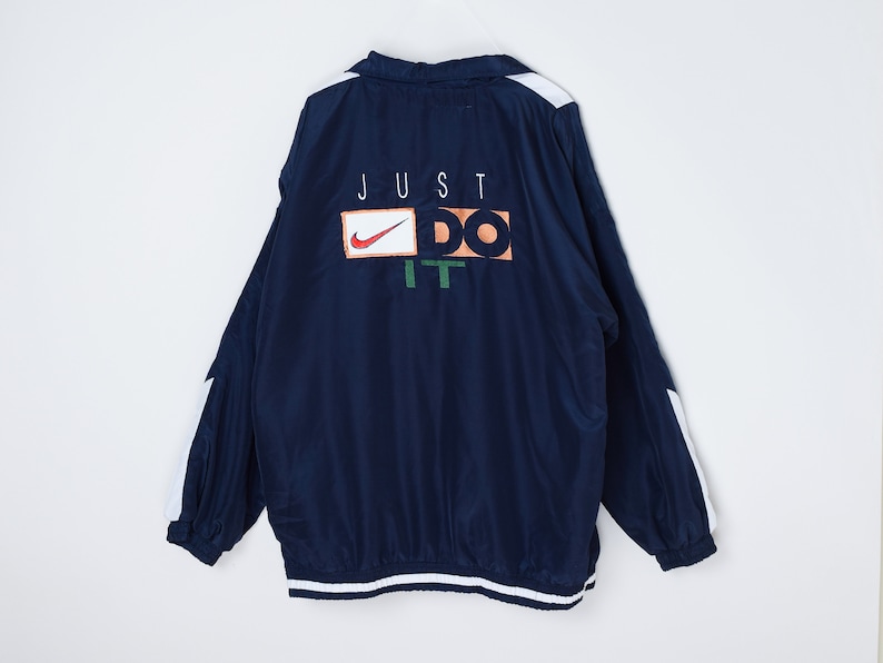 May include: A navy blue windbreaker jacket with white accents on the sleeves and hem. The back of the jacket features a white "JUST DO IT" logo with a Nike swoosh.