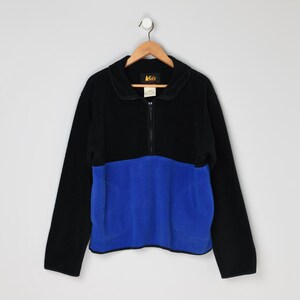 May include: Black and blue fleece pullover with a half-zip closure. The top portion is black, and the lower section is a vibrant blue. The garment is hanging on a wooden hanger against a plain white background.