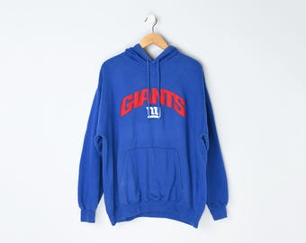 May include: A blue hooded sweatshirt with the word "GIANTS" in red block letters across the chest. The sweatshirt has a front pocket and a drawstring hood. The garment is hanging on a wooden hanger.