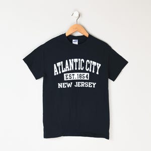 Può includere: Una maglietta nera con testo bianco che recita "Atlantic City Est. 1854 New Jersey".