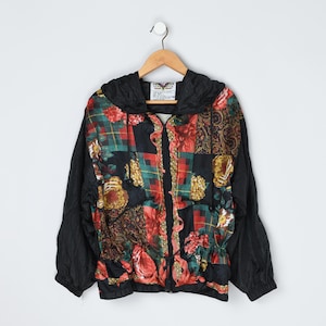 May include: A vintage black jacket with a patchwork design featuring floral, plaid, and paisley patterns in red, green, and gold. The jacket has a hood and black sleeves, hanging on a wooden hanger against a white background.