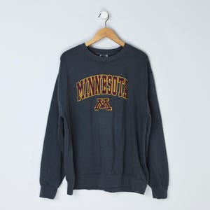 May include: A dark gray sweatshirt with the word "MINNESOTA" arched across the chest in gold and maroon lettering. Below the word is a maroon "M" logo. The sweatshirt is hanging on a wooden hanger against a white background.