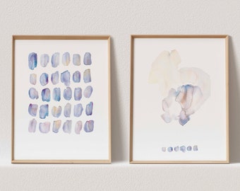 Puede incluir: Dos pinturas de acuarela enmarcadas con diseños abstractos en tonos azules, morados y beige. La pintura de la izquierda presenta una cuadrícula de pequeñas formas irregulares. La pintura de la derecha presenta una sola forma grande e irregular.