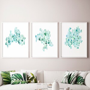 May include: Three framed abstract watercolor paintings with a teal and blue color scheme. The paintings are hung on a light pink wall above a white sofa with green leaf patterned throw pillows.
