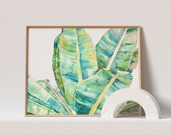 Op de afbeelding: Een rechthoekige ingelijste kunstdruk met een aquarel van grote, levendige groene en blauwgroene bananenbladeren met zichtbare nerven. Een klein, wit, halfrond stenen boogsculptuur staat voor de rechterbenedenhoek van het kunstwerk.