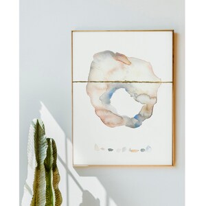 May include: Abstract watercolor painting in a gold frame. The artwork features a central, organic shape in muted tones of peach, gray, and blue, bisected by a gold line. A collection of small, colorful shapes is below the main form.