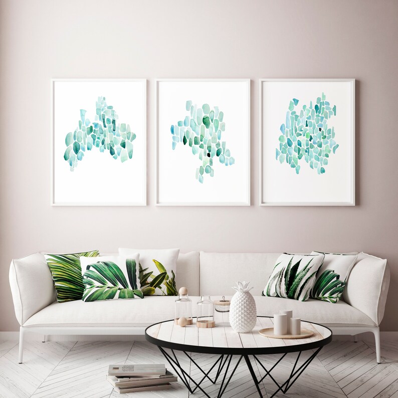 May include: Three framed abstract watercolor paintings with a teal and blue color scheme. The paintings are hung on a light pink wall above a white sofa with green leaf patterned throw pillows.