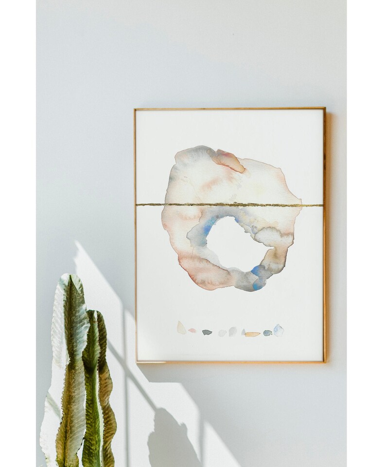 May include: Abstract watercolor painting in a gold frame. The artwork features a central, organic shape in muted tones of peach, gray, and blue, bisected by a gold line. A collection of small, colorful shapes is below the main form.