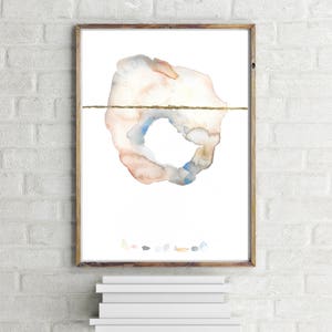 May include: Abstract watercolor art print in a wooden frame. The artwork features a circular shape with a gold horizontal line, and a palette of soft colors including peach, blue, and gray. The print is displayed on a white background.
