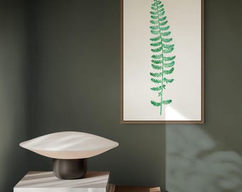 Op de afbeelding: Een ingelijste verticale wandkunstprint met een illustratie van een groene varenblad op een crèmekleurige achtergrond, naast een paddenstoelvormige tafellamp met een witte glazen kap en een kleine zwarte kom op een stapel boeken.