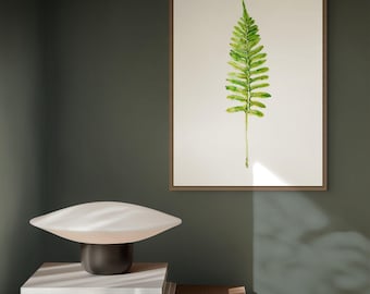 Op de afbeelding: Een ingelijste wandkunstprint met een illustratie van een enkele groene varenblad op een crèmekleurige achtergrond. Daaronder staat een paddenstoelvormige tafellamp met een witte glazen kap en een donkere voet naast een zwarte kom.