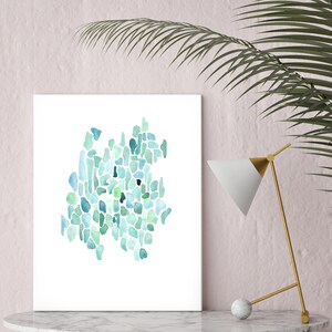 May include: A framed abstract watercolor painting with a teal, blue, and green color scheme. The painting is on a white background and is displayed on a white marble table with a gold lamp.