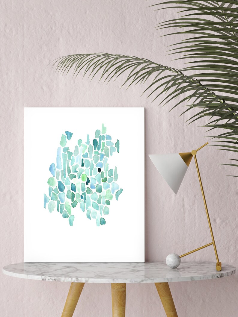 May include: A framed abstract watercolor painting with a teal, blue, and green color scheme. The painting is on a white background and is displayed on a white marble table with a gold lamp.