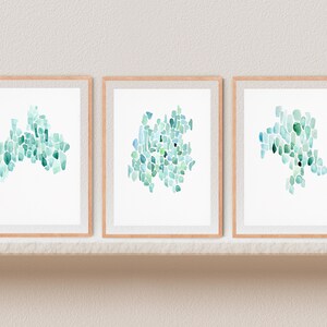 May include: Three framed watercolor paintings with abstract designs in shades of blue and green. The paintings are displayed on a white shelf against a white wall.