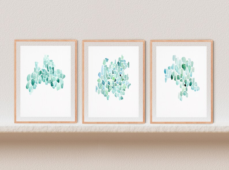 May include: Three framed watercolor paintings with abstract designs in shades of blue and green. The paintings are displayed on a white shelf against a white wall.