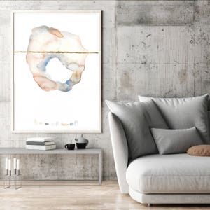 May include: Abstract watercolor art print in a white frame. The artwork features a circular shape with peach, blue, and gray tones, bisected by a gold line. The print is displayed above a gray table with books, a teapot, and a coffee cup.