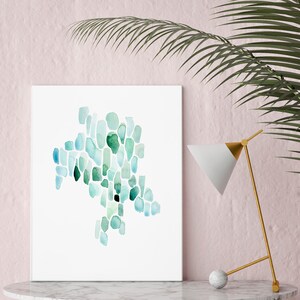 May include: A framed abstract watercolor painting with a teal and green color scheme. The painting is on a white background and is displayed on a white marble table with a gold lamp.