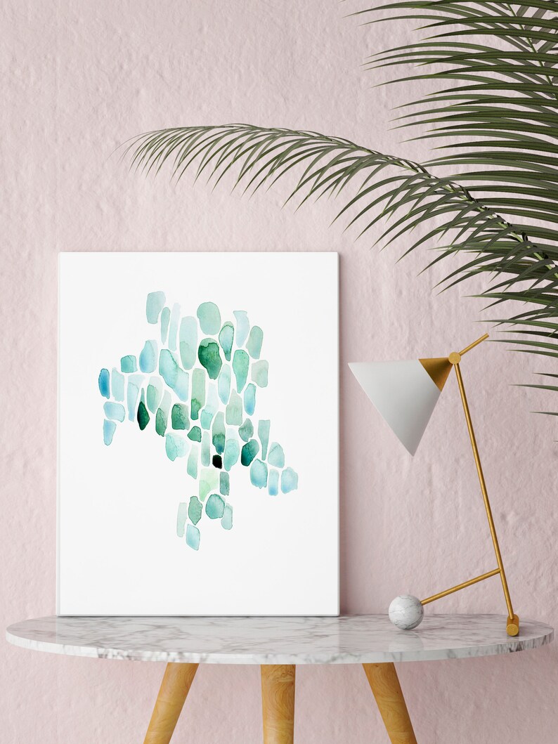 May include: A framed abstract watercolor painting with a teal and green color scheme. The painting is on a white background and is displayed on a white marble table with a gold lamp.