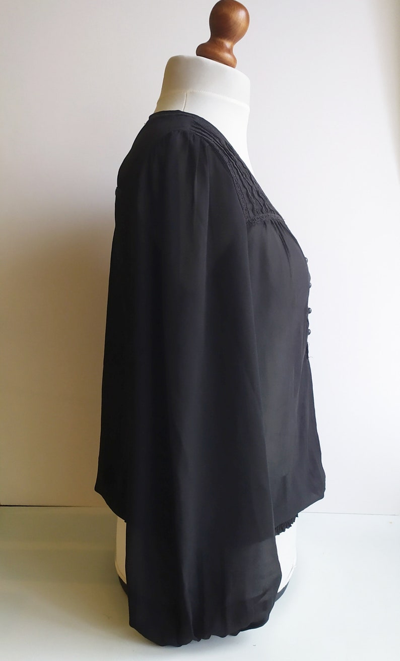 May include: A black sheer blouse with a lace trim around the neckline and a gathered elastic band at the bottom of the sleeves. The blouse has a v-neckline and a button closure at the front.