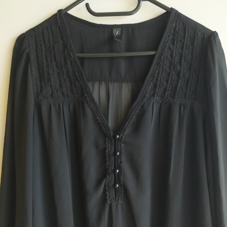 May include: A black sheer blouse with a lace trim around the neckline and a button closure. The blouse has long sleeves and a v-neck.
