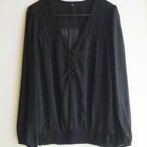 May include: A black sheer blouse with a lace trim around the neckline and a button detail down the front. The blouse has long sleeves and a gathered elastic waistband.