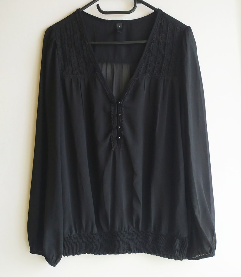 May include: A black sheer blouse with a lace trim around the neckline and a button detail down the front. The blouse has long sleeves and a gathered elastic waistband.