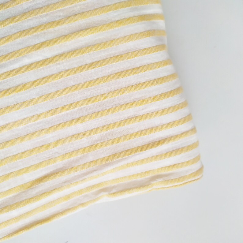 May include: Yellow and white striped fabric with a subtle texture. The fabric is folded over on itself, showing the underside of the fabric.