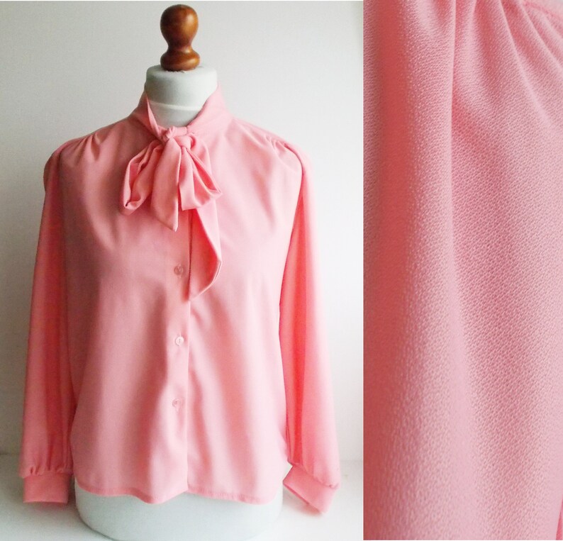 May include: A pink long-sleeved blouse with a large bow at the neck. The blouse has a button-up front and a slightly puffed sleeve.