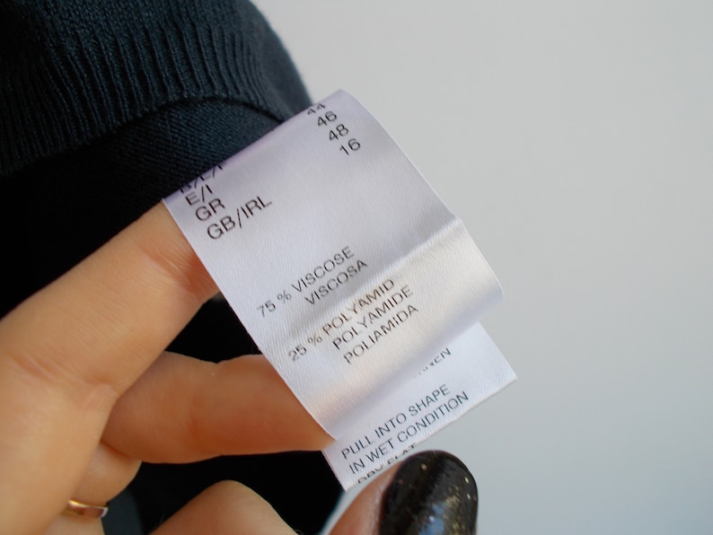 K&ouml;nnte beinhalten: Wei&szlig;es Kleidungsetikett mit schwarzem Text. Das Etikett lautet "75% Viskose Viscosa 25% Polyamid Polyamide Poliamida". Das Etikett lautet auch "Pull into shape in wet condition".