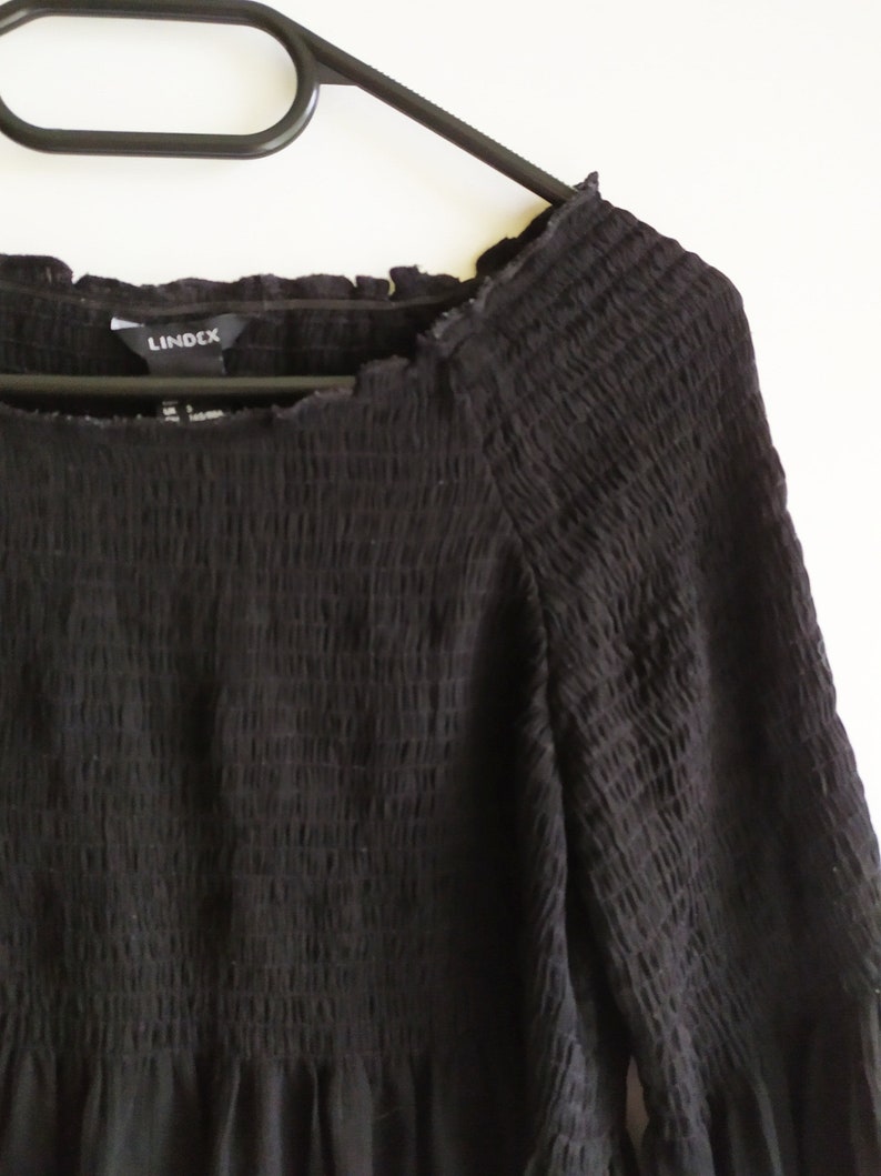 May include: Black, long-sleeved, off-the-shoulder dress with a smocked bodice and a ruffled hem. The dress has a "Lindex" brand label.