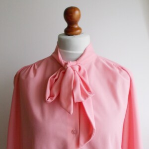 May include: A pink blouse with a large bow tie at the neck. The blouse has long sleeves and a button-up front.